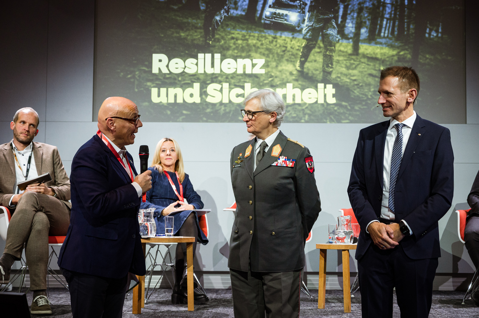 <br /> Mann auf Bühne spricht in Handmikrofon. Drei weitere Männer und eine Dame sind ebenfalls auf der Bühne.